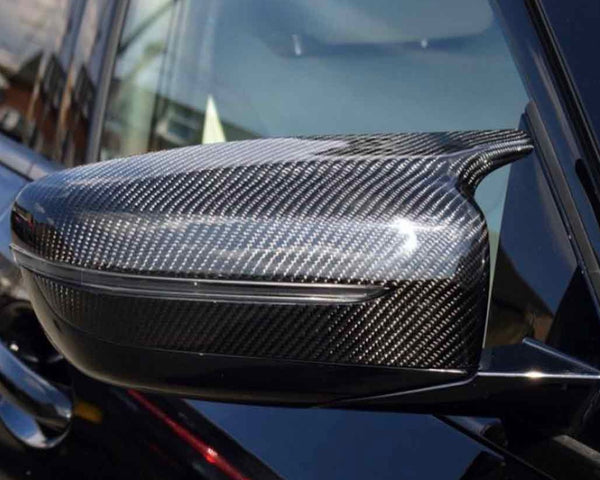 A carbon fiber car side mirror, reflecting light, is attached to the sleek black exterior of a vehicle, with a blurred urban background visible.
