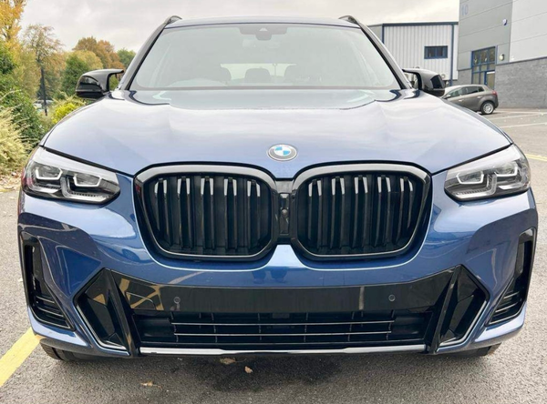 A blue BMW SUV is parked in a parking lot, displaying a prominent front grille and headlights, with trees and industrial buildings in the background.