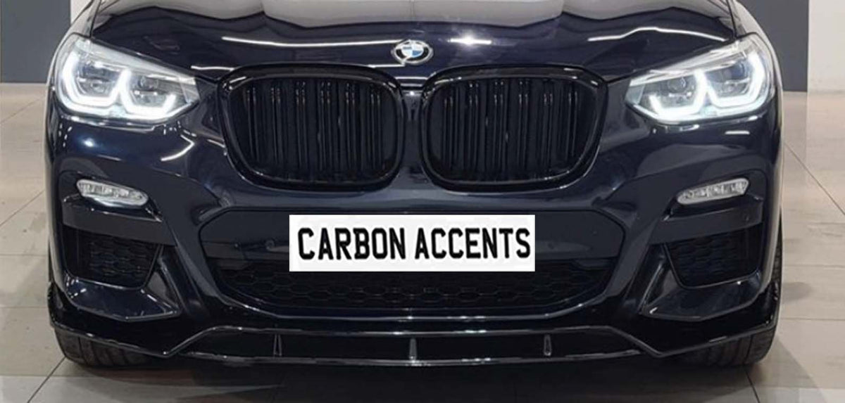 Car front view displaying illuminated headlights in a showroom setting. Prominently featured below the grille is a sign reading "CARBON ACCENTS."