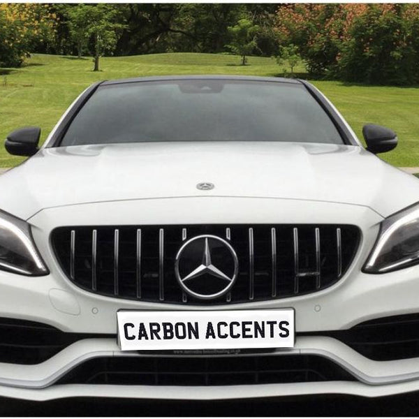 A white car is parked on a driveway, displaying the text "CARBON ACCENTS" on its front plate. The background features a garden with grass and trees.