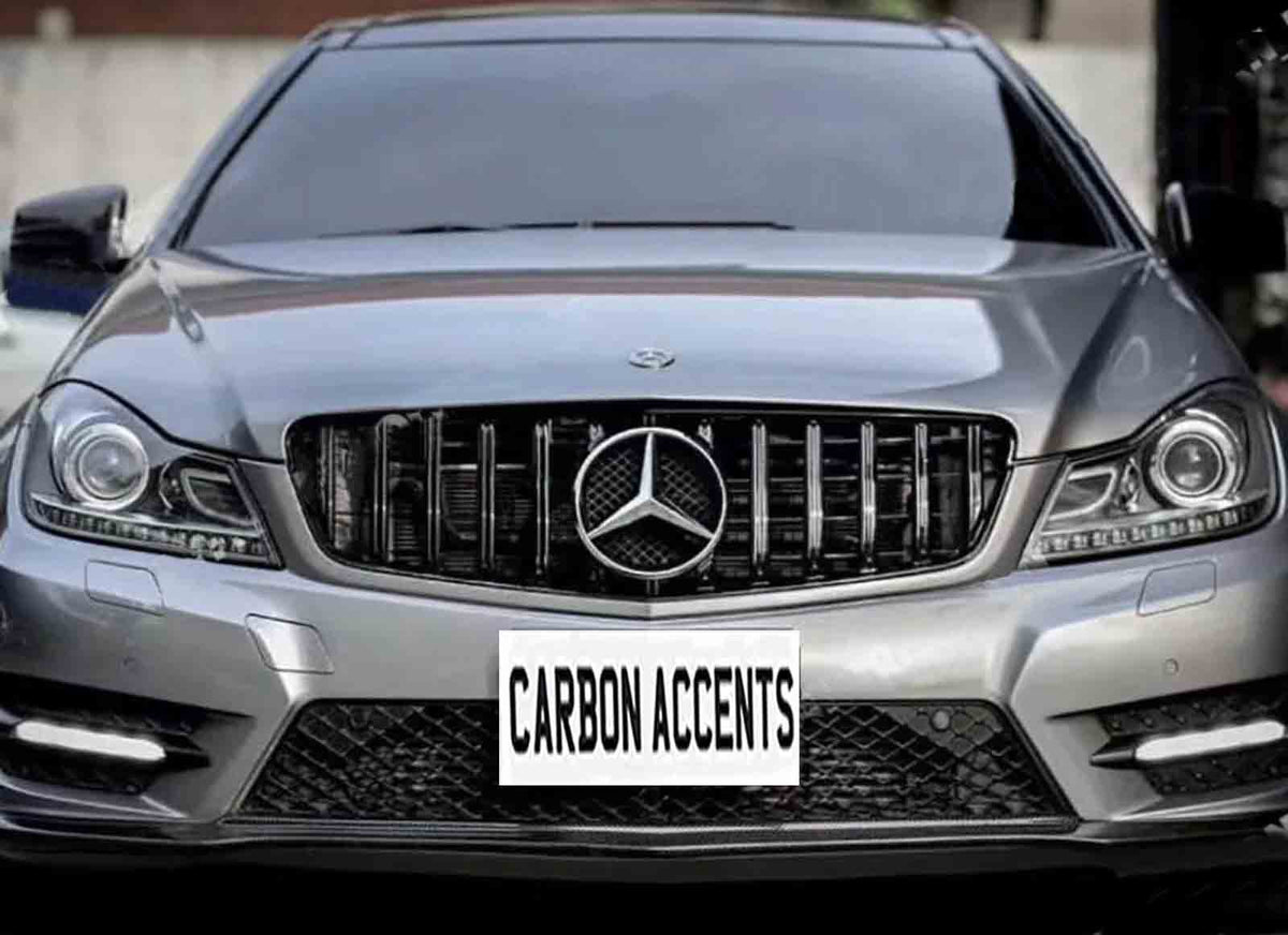 A silver car features a front grille with a central logo, parked in an urban area. The license plate reads "CARBON ACCENTS."