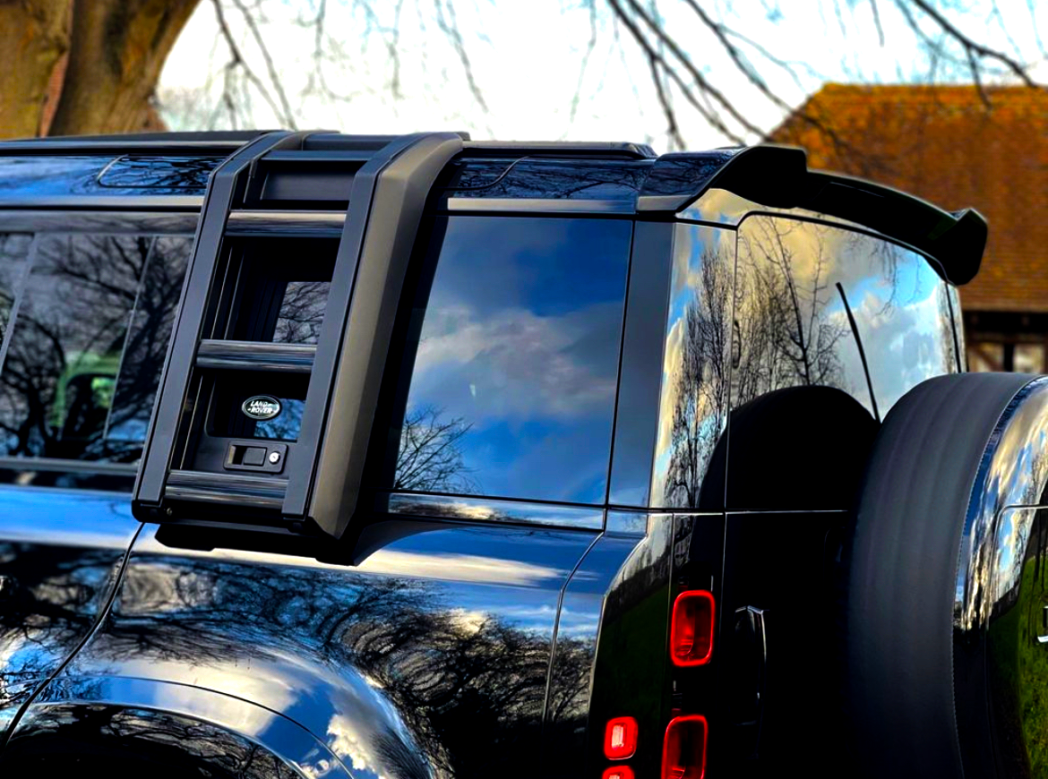 A black off-road vehicle with a mounted ladder and spare tire, reflecting trees and sky, is parked near a tree. A logo reads "Land Rover."