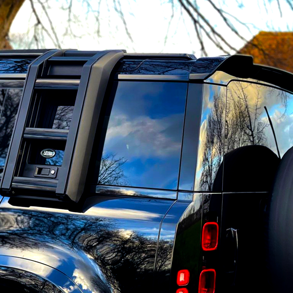 A black off-road vehicle with a mounted ladder and spare tire, reflecting trees and sky, is parked near a tree. A logo reads "Land Rover."