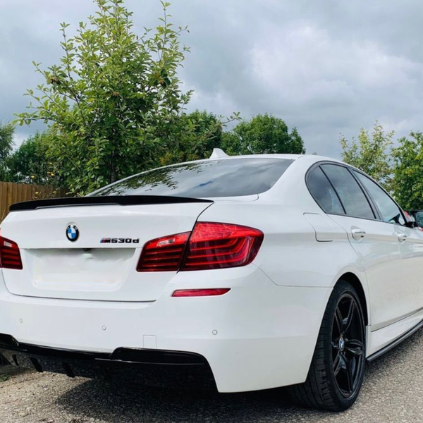 A white BMW 530d car is parked on a street. It has black wheels and red taillights, with trees and cloudy skies in the background.