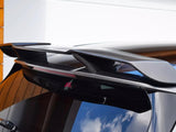 A carbon fiber car spoiler is mounted on the roof of a black vehicle, parked against a wooden structure and white wall, with blue sky reflections visible on the window.