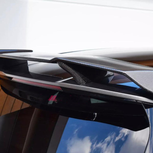 A carbon fiber car spoiler is mounted on the roof of a black vehicle, parked against a wooden structure and white wall, with blue sky reflections visible on the window.