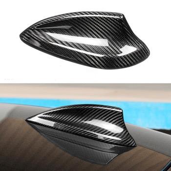 A carbon fiber shark fin antenna rests on a car roof, reflecting light. The surface around it shows a sky-blue hue, suggesting outdoor daylight conditions.