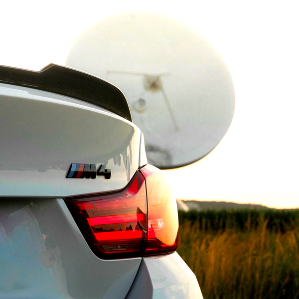A white car with "M4" emblem and illuminated tail light is parked, partially obscuring a large satellite dish in a grassy field at sunset.
