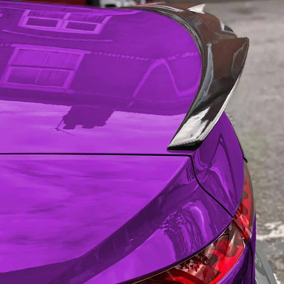 A vibrant purple car with a carbon fiber spoiler is parked, reflecting building windows on its glossy surface. Tail lights are partially visible, hinting at a rear view.