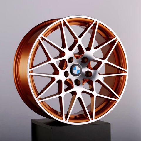 A bronze alloy wheel with intricate spoke design and a central blue and white emblem, resting on a black pedestal against a neutral gray background.