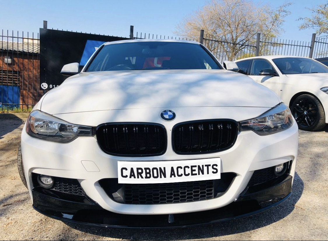 A white BMW sedan is parked outdoors, facing forward. The license plate reads "CARBON ACCENTS." Other cars and a fence are visible in the background.
