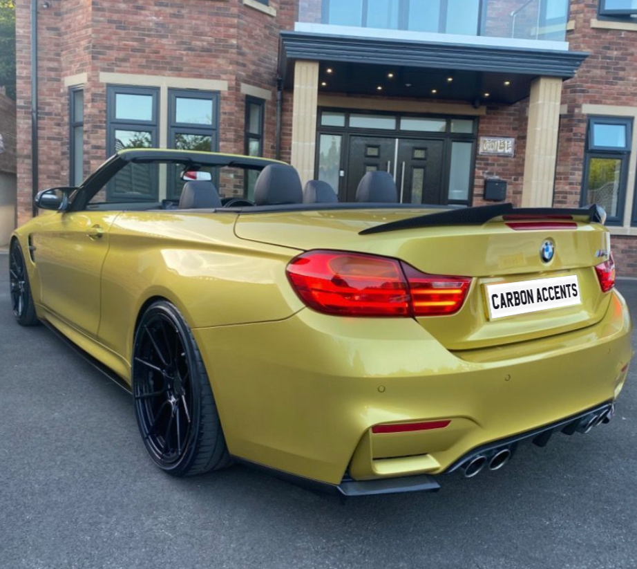 A gold convertible sports car with a visible spoiler is parked in front of a brick building. The license plate reads "CARBON ACCENTS."