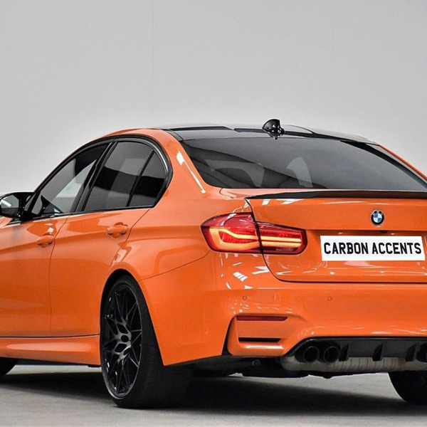 Orange BMW M3, parked, features "CARBON ACCENTS" on its rear plate, displayed in a bright showroom with a grey and white wall background.