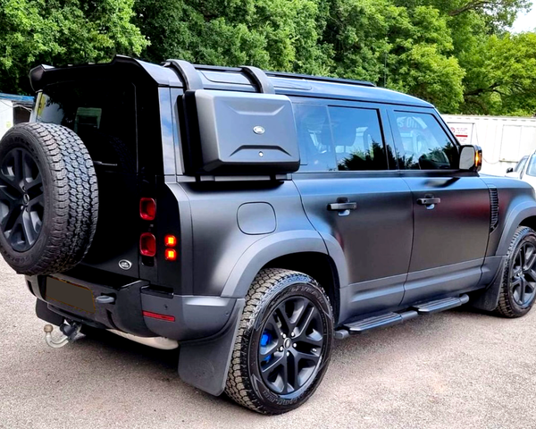 A black off-road SUV is parked on a paved area, featuring a roof rack, spare tire, and side storage box, surrounded by trees and vehicles in the background.