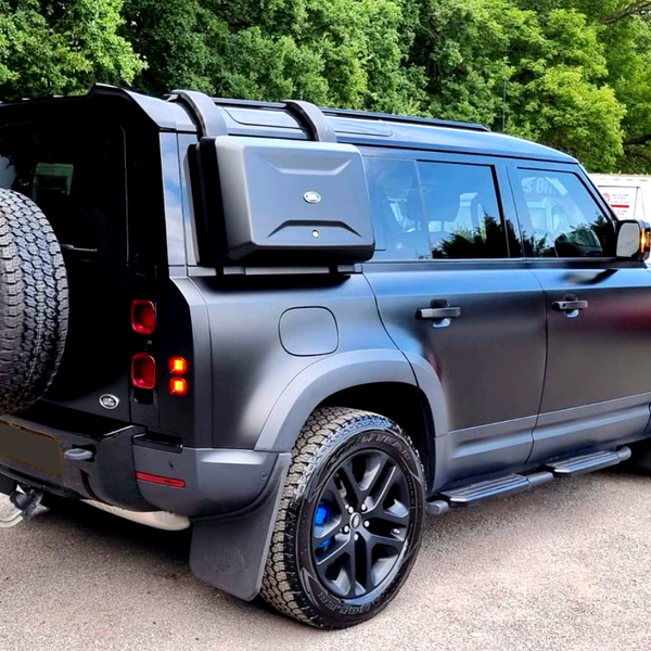 A black off-road SUV is parked on a paved area, featuring a roof rack, spare tire, and side storage box, surrounded by trees and vehicles in the background.