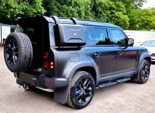 A black off-road SUV is parked on a paved area, featuring a roof rack, spare tire, and side storage box, surrounded by trees and vehicles in the background.