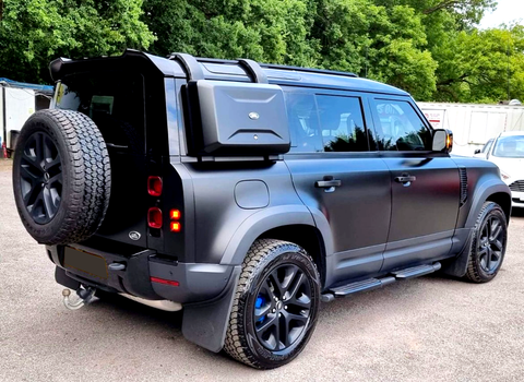 A black off-road SUV is parked on a paved area, featuring a roof rack, spare tire, and side storage box, surrounded by trees and vehicles in the background.