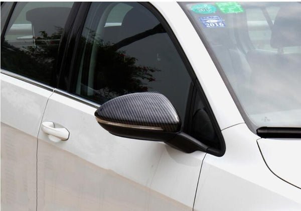 A white car features a carbon-fiber side mirror reflecting nearby trees, parked in an outdoor setting. Stickers, including a 2016 sticker, are displayed on the windshield.