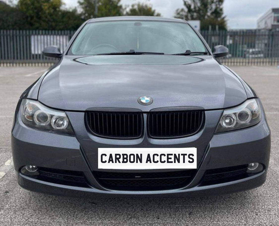 A gray BMW sedan is parked in a lot, viewed from the front. The license plate reads "CARBON ACCENTS," with a fence and trees in the background.