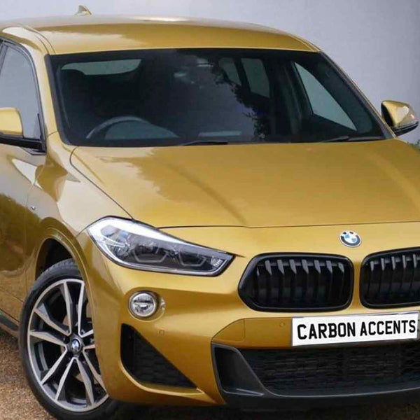 A gold BMW parked on a gravel driveway, with a white wall and greenery in the background. The license plate reads "CARBON ACCENTS."