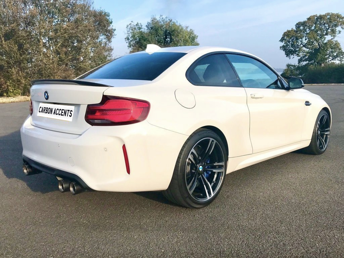 A white sports car, parked on an asphalt surface, features shiny black rims and dual exhaust. Trees and a clear sky are visible in the background. The license plate reads, "CARBON ACCENTS."