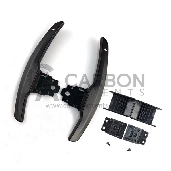 Curved paddle shifters with "+" and "-" symbols, accompanied by brackets and screws, are laid out on a white background. Text: "CARBON ACCENTS www.carbonaccents.com".