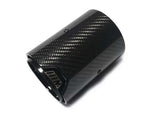 A carbon fiber exhaust tip lies against a plain white background. The surface has a textured pattern. The inner edge features a logo or marking resembling 