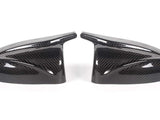 Two carbon fiber side mirror covers rest on a white surface, showcasing a glossy finish and intricate weave pattern, appearing symmetrically aligned.