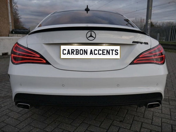 A white luxury car is stationary in a parking area. The rear features sleek taillights and dual exhausts. The license plate reads "CARBON ACCENTS" and skies are overcast.