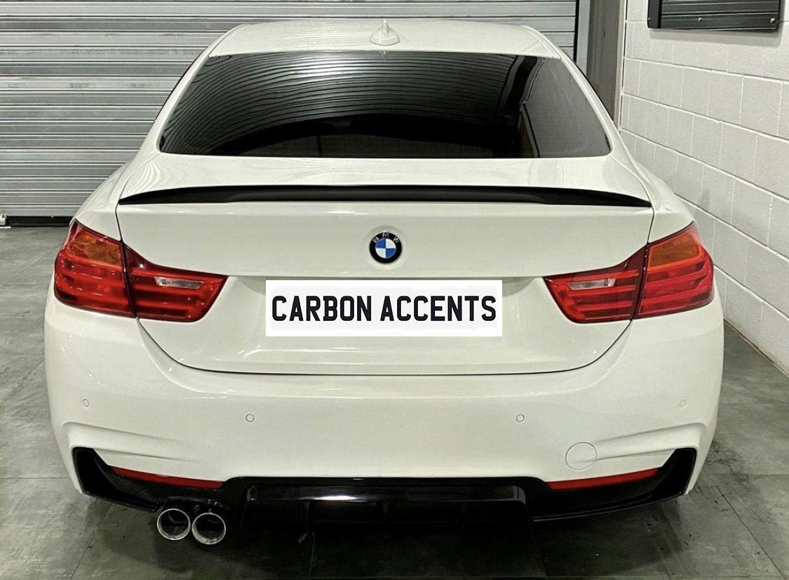 White BMW sedan with tinted windows and dual exhaust pipes is parked in a garage. The license plate reads "CARBON ACCENTS." A metallic wall and concrete floor surround the car.