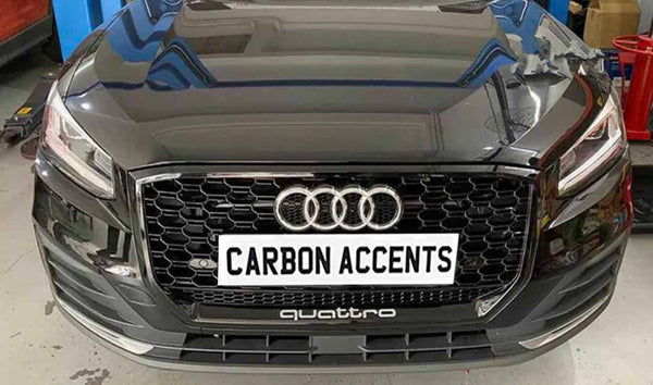 A black car's front view with a honeycomb grille and prominent logo, displaying the text "CARBON ACCENTS" on a plate, set in a garage environment with tools around.