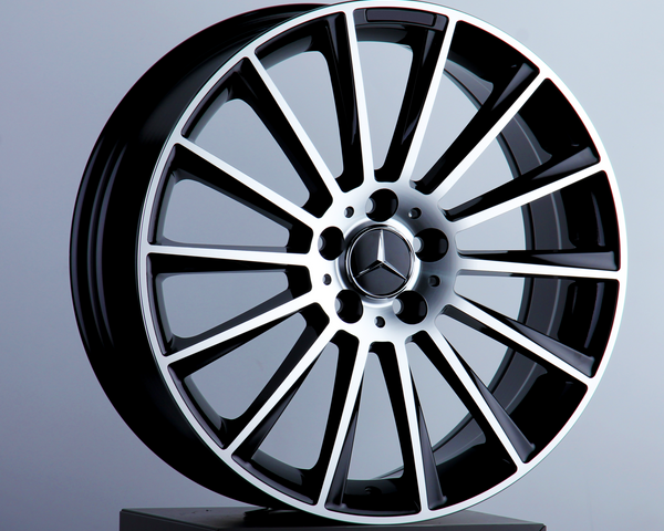 A glossy, black and chrome alloy wheel rests on a pedestal, featuring a three-pointed star emblem at its center, set against a neutral gray background.