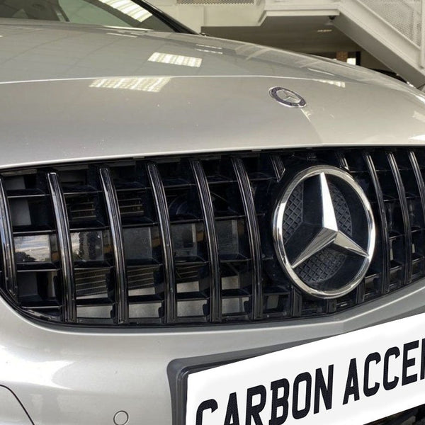 The silver car grille displays a prominent three-pointed logo, surrounded by vertical black slats in a showroom. Below, a white license plate reads "CARBON ACCENTS."