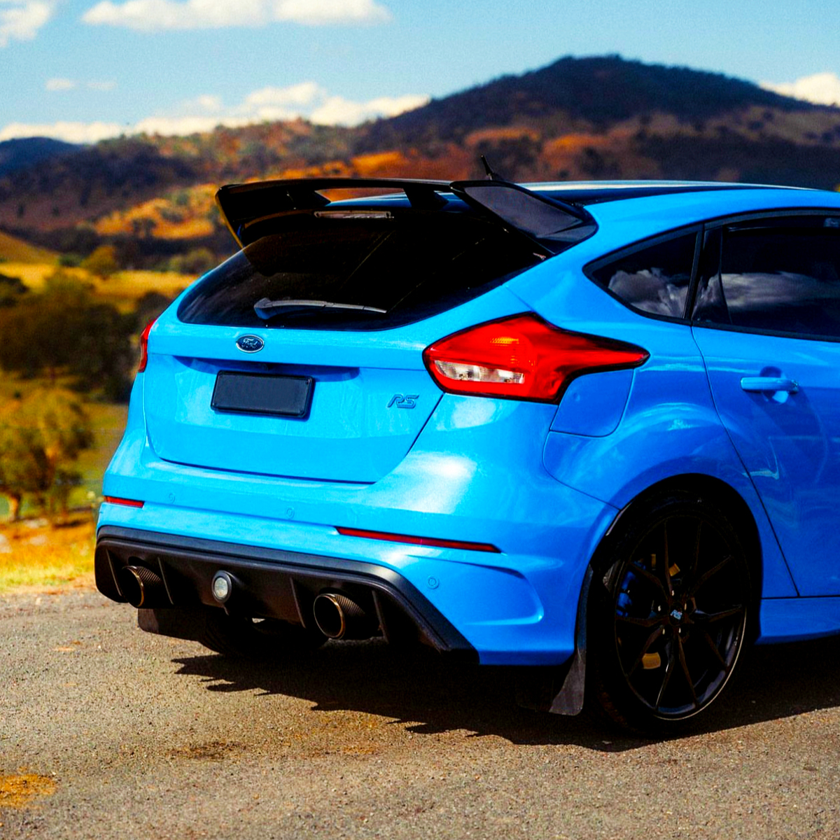 A vibrant blue sports car is parked on a roadside, featuring dual exhausts and a prominent spoiler, with scenic hills and a clear blue sky in the background.