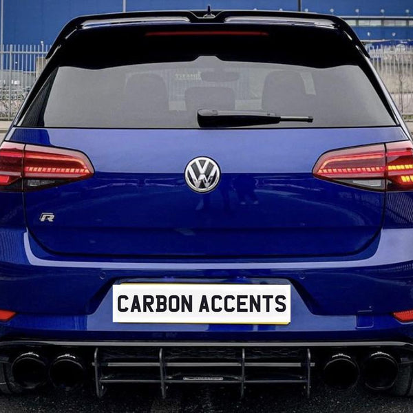 A blue Volkswagen car is parked, displaying its rear with red taillights illuminated. The license plate reads "CARBON ACCENTS," surrounded by an urban parking area with metal fencing.