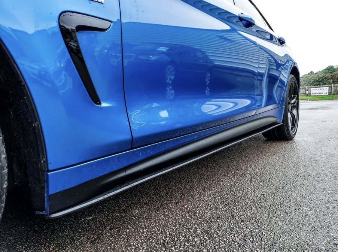 Blue car parked on wet pavement, reflecting the surroundings. A black trim accentuates the side, with “BMW” visible. Background shows greenery and a sign reading “CHEVEN PARKING.”
