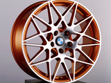 The bronze alloy wheel features an intricate, multi-spoke design, resting on a black pedestal. A blue-and-white circular emblem is at the center, set against a plain background.