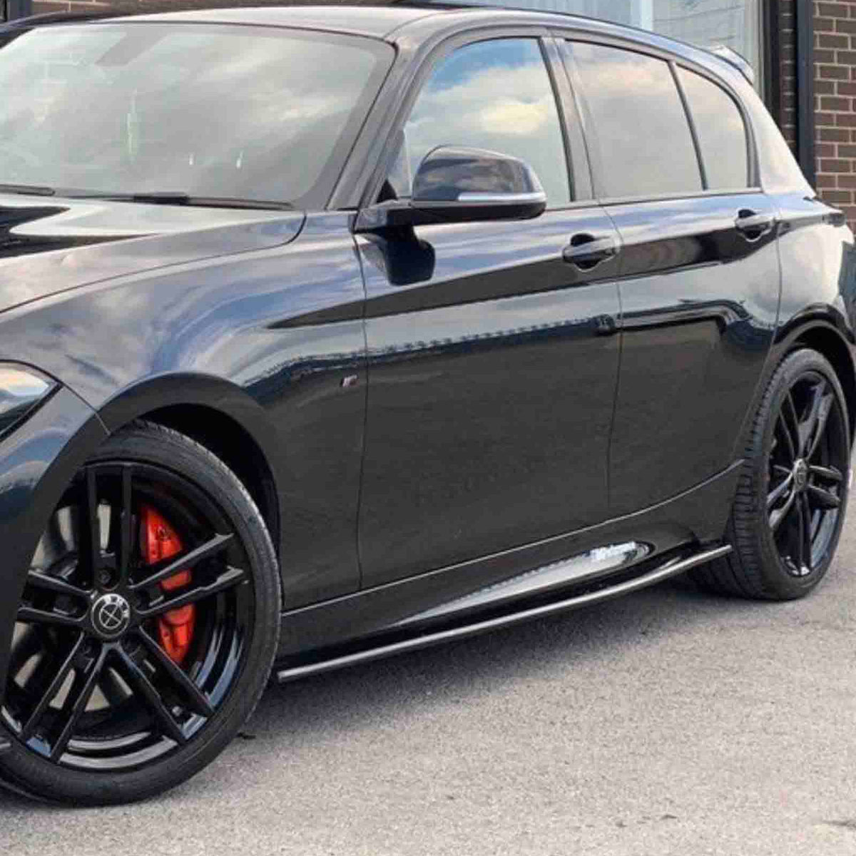 A black car with red brake calipers is parked on a paved surface, alongside a brick building with large windows.