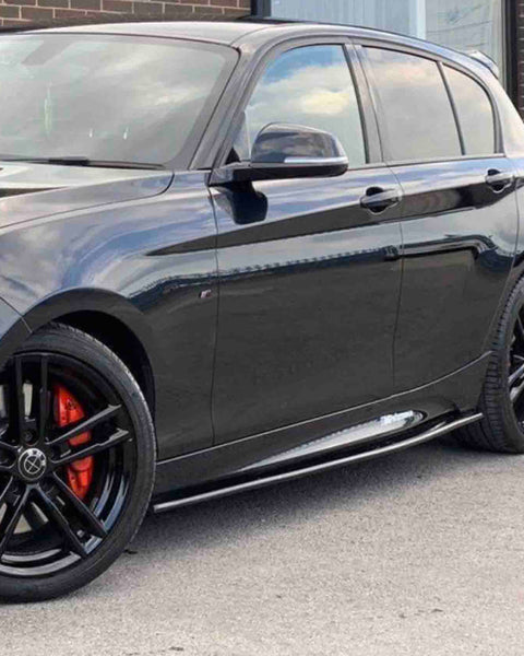 A black car with red brake calipers is parked on a paved surface, alongside a brick building with large windows.