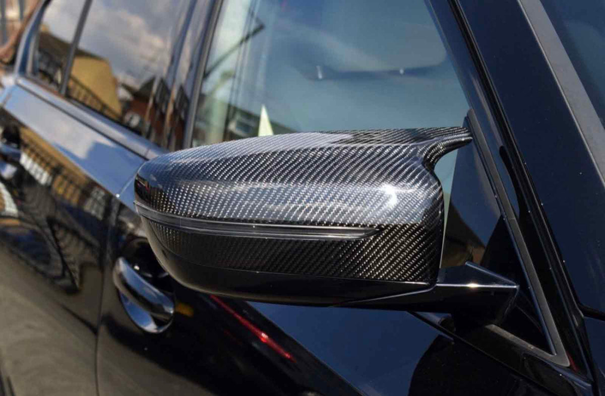 Carbon fiber car side mirror reflecting the sunlight, attached to a sleek black vehicle. The background shows blurred buildings and a clear sky.