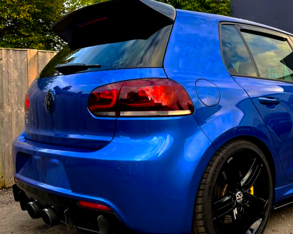 A blue car with tinted taillights and a prominent spoiler is parked on a gravel surface, next to a wooden fence and greenery, under a partly cloudy sky.