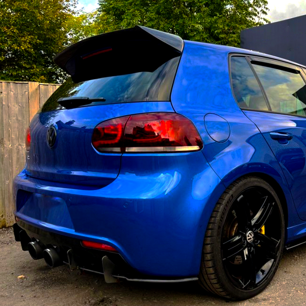 A blue car with tinted taillights and a prominent spoiler is parked on a gravel surface, next to a wooden fence and greenery, under a partly cloudy sky.