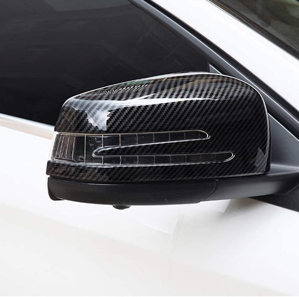 A car's side mirror with a carbon fiber design cover, featuring integrated indicator lights, mounted on a white vehicle in an outdoor setting.