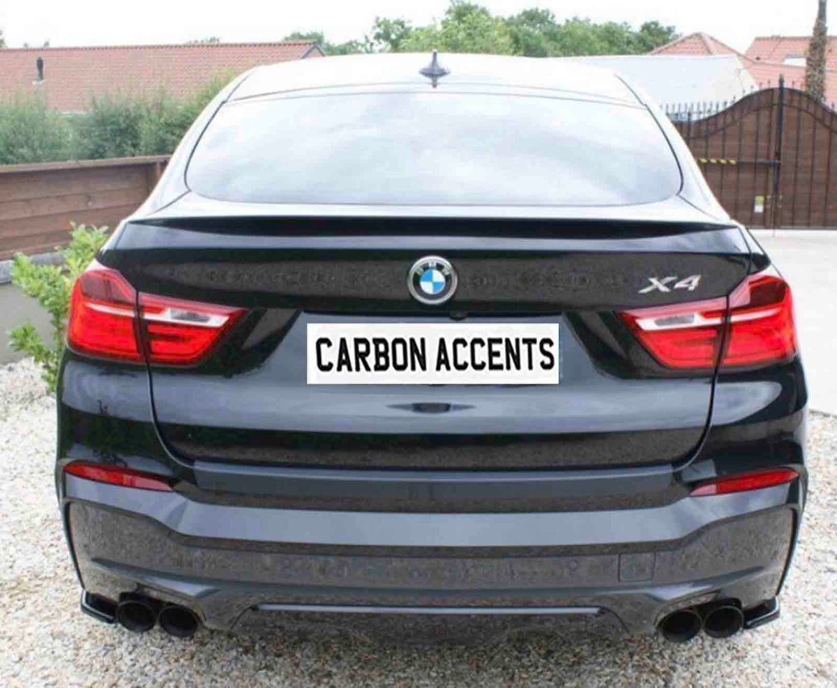 A black BMW X4 with visible taillights is parked on gravel. The license plate reads "CARBON ACCENTS." Trees and a fence form the background environment.