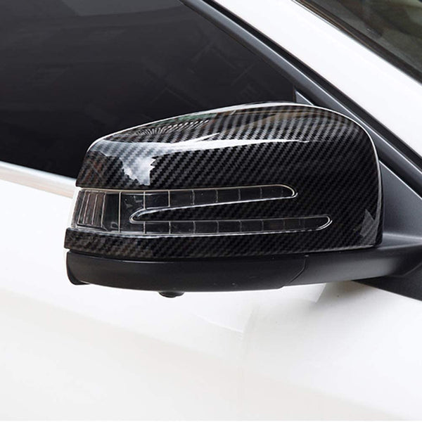 A carbon fiber side mirror cover on a white car, displaying a sleek design with integrated indicator lights, positioned against a dark window background.