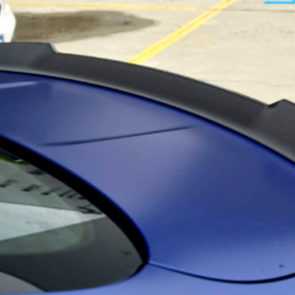A blue car features a black spoiler, parked on a concrete surface. Another white car is partially visible in the background, highlighting a parking lot setting.