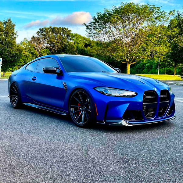A blue sports car is parked on an asphalt surface, surrounded by lush green trees under a partly cloudy sky.