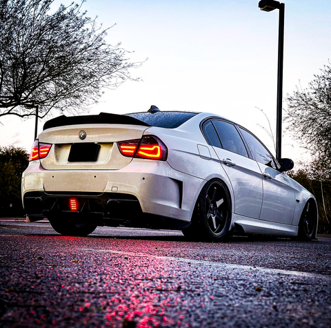 A white car with illuminated taillights is parked on a wet road, reflecting red light. Surrounding it are street lamps and leafless trees against a clear sky.