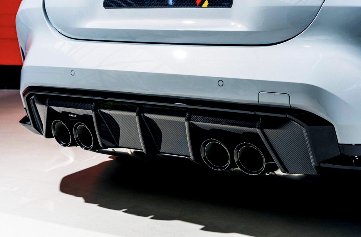 A white car features quad exhaust pipes integrated into a black carbon fiber diffuser, displayed in a well-lit indoor setting with a red wall in the background.