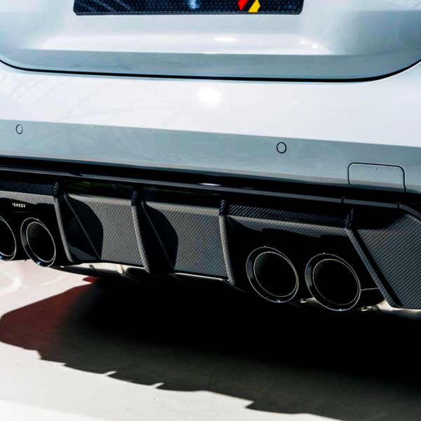 A white car features quad exhaust pipes integrated into a black carbon fiber diffuser, displayed in a well-lit indoor setting with a red wall in the background.